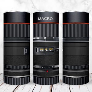 以下が含まれることがあります： 黒と銀のカメラレンズのデザインが施されたステンレス製のタンブラー。レンズには、フィートとメートルでマークされた被写界深度スケールがあります。タンブラーには「MACRO」という文字が印刷されています。