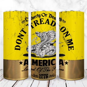 Puede incluir: Un vaso amarillo con un acabado envejecido, con la frase "Don't Tread on Me" y una serpiente enroscada. La sección inferior es de color dorado metálico, con las palabras "America, Land of the Free, 1776".