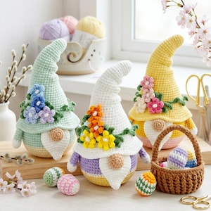 Puede incluir: Tres gnomos de ganchillo con sombreros altos y curvos y adornos florales. Los gnomos son de colores pastel: verde, blanco y amarillo. Una pequeña cesta contiene huevos de Pascua de ganchillo. Hay huevos de ganchillo adicionales esparcidos alrededor de los gnomos. El tema es la Pascua y la primavera.