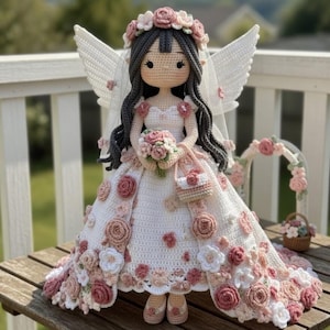 May include: A handmade crochet doll in a bridal gown, featuring a floral crown, wings, and a dress decorated with pink and white flowers. The doll holds a bouquet and a small purse, displayed on a wooden table.