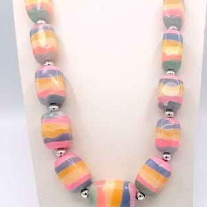 May include: A colorful necklace featuring large, barrel-shaped beads. The beads are striped with shades of pink, yellow, blue, and green, separated by silver-colored metal beads. The necklace is displayed on a white stand.