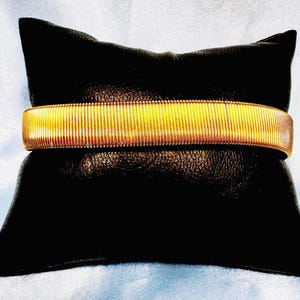 Puede incluir: Un brazalete dorado extensible con textura acanalada. El brazalete se muestra sobre un cojín de terciopelo negro.