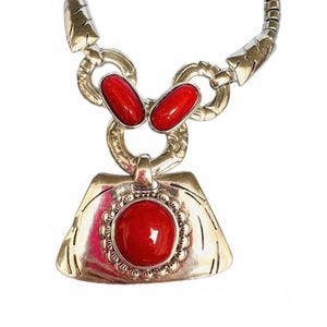 May include: Silver-toned necklace featuring a pendant with a large, round red stone surrounded by a decorative border. Two smaller red stones are set above the pendant. The necklace has a chain-like design.