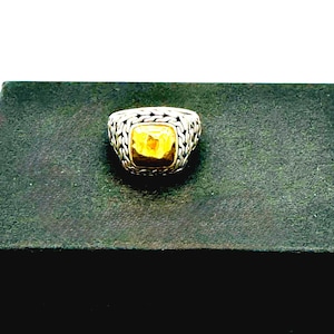 Pode incluir: Um anel de prata com uma pedra preciosa amarela quadrada. O anel tem um design tecido em volta da pedra preciosa. O anel está sobre uma superfície verde escura.