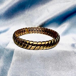 Op de afbeelding: Een goudkleurige, rekbare armband met een geribbeld ontwerp. De armband is gemaakt van meerdere rechthoekige segmenten die met elkaar verbonden zijn, waardoor een flexibele band ontstaat. Het oppervlak heeft een getextureerd, geribbeld patroon dat licht reflecteert. De achtergrond is een lichtblauwe stof.