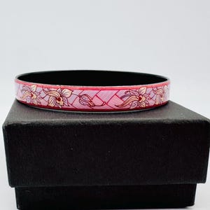 May include: A pink and black bangle bracelet with a floral design. The bracelet features a repeating pattern of flowers in shades of pink, white, and beige. The interior of the bracelet is black.
