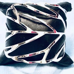 May include: A silver metal cuff bracelet with an open, geometric design. The bracelet is displayed on a black, textured surface. The cuff's design features interconnected, curved lines, creating an eye-catching accessory.
