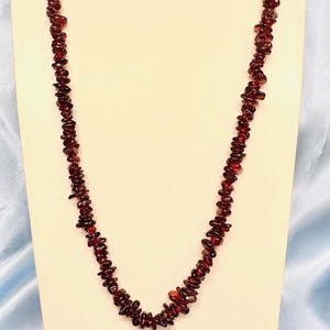 May include: A necklace and bracelet set featuring small, irregularly shaped, dark red gemstones. The jewellery is displayed on a cream-coloured bust against a light blue backdrop. The necklace hangs in a U-shape.