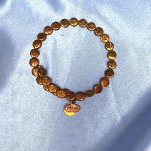 May include: A gold-colored bracelet composed of small, round, textured beads. A small, oval charm with the text "VINTAGE SIXTY-SIX" hangs from the bracelet. The bracelet is displayed on a light blue, satin-like fabric.