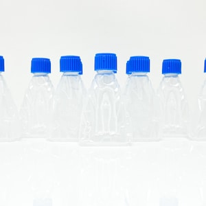 Puede incluir: Colección de siete botellas de plástico transparente con tapas azules de rosca. Las botellas tienen forma triangular y están dispuestas en fila sobre un fondo blanco. Las tapas son de un azul vibrante.