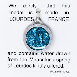 May include: A round, silver-toned medal with a blue center depicting a religious scene. The medal is inscribed with the words "LOURDES" and "FRANCE". The text on the card states the medal contains water from the Miraculous spring of Lourdes.