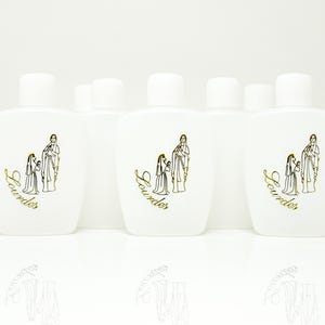 May include: White, oval-shaped bottles with white caps. Each bottle features a gold illustration of two figures and the word "Lourdes" in gold script. The bottles are arranged in a row, reflecting on a white surface.