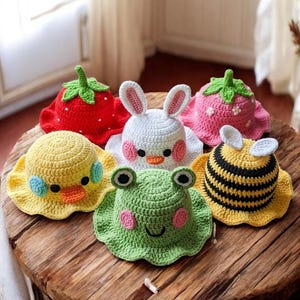May include: A collection of six handmade crochet hats in various animal and fruit designs. The hats include a red strawberry, a pink strawberry, a white bunny, a yellow duck, a green frog, and a yellow bee. The hats are displayed on a wooden surface.