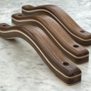 May include: Three wooden cabinet handles with a curved design. The handles are made of dark brown wood with a light-colored wood accent. Each handle has a hole at each end for mounting. The handles are arranged in a row, slightly overlapping.