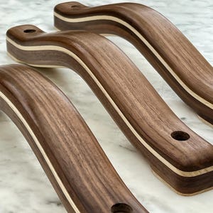 May include: Three curved wooden handles with a dark brown finish and a light-colored stripe. Each handle has a rounded shape with a hole drilled at each end. The handles are likely for furniture or cabinetry.