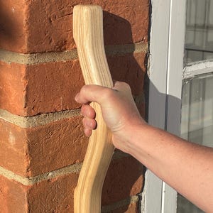 Puede incluir: Un mango de madera clara y curvada. El mango se sostiene contra una pared de ladrillos rojos. La veta de la madera es visible. El mango es liso y parece estar diseñado ergonómicamente.