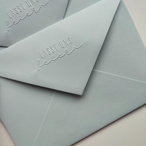 Könnte beinhalten: Hellblaue Umschläge mit dem geprägten Text „LASST UNS feiern“. Die Umschläge sind in einer geschichteten Komposition angeordnet, wobei die Klappe des oberen Umschlags geöffnet ist. Das Papier hat eine strukturierte Oberfläche.
