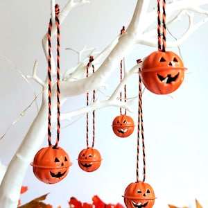 May include: Orange metal jack-o'-lantern decorations suspended from a white tree. Each pumpkin has a carved face and is hung by a black and orange twisted cord. A large pumpkin and autumn leaves are at the base.