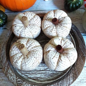 May include: Four fabric pumpkins with cream-coloured fabric and script print, arranged in a wooden tray. Each pumpkin has a brown stem and is a decorative autumn accent. Other pumpkins are in the background.