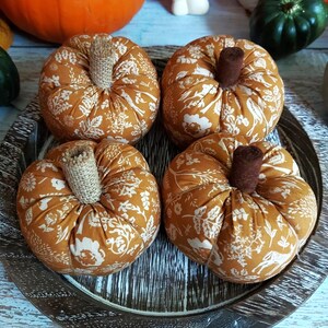 May include: Four fabric pumpkins with tan and white floral patterns sit on a wooden tray. Each pumpkin has a brown stem. The pumpkins are a warm orange colour, perfect for autumn decor.