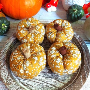 May include: Four fabric pumpkins with a mustard yellow background and a white floral pattern, each featuring a textured stem. These decorative pumpkins are arranged on a round, dark brown wooden tray with a visible grain and distressed finish.