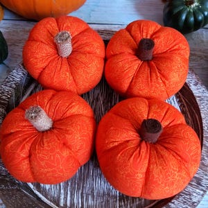 May include: Four orange fabric pumpkins with brown stems on a wooden tray. The pumpkins have a floral pattern and are arranged in a square. The background includes a larger orange pumpkin and decorative items.