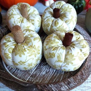 May include: Four fabric pumpkins with white and gold floral patterns and brown stems sit on a wooden tray. The pumpkins are round and have a textured appearance, ideal for autumn decorations.