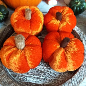 May include: Four plush, orange velvet pumpkins with brown and tan stems sit on a decorative wooden tray. The pumpkins are arranged in a circular pattern, with a larger pumpkin and other autumn decorations in the background.