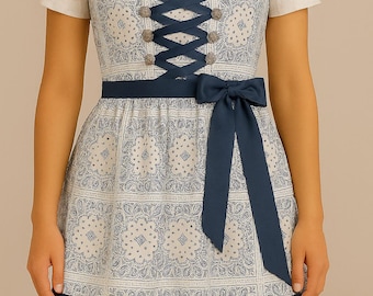 May include: A traditional dirndl dress with a white bodice and short sleeves. The dress features a blue and white paisley pattern, a navy blue ribbon corset, and a matching bow at the waist. The skirt has navy blue trim.
