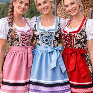 Op de afbeelding: Drie vrouwen in traditionele Duitse dirndl-jurken. De jurken hebben bloemenlijfjes, gesmokte korsetten en kleurrijke schorten. De schorten zijn roze, blauw en rood, met decoratieve randen. De vrouwen lachen en poseren voor de camera.