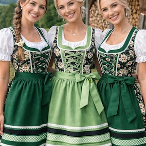 Op de afbeelding: Drie vrouwen in traditionele Duitse dirndl-jurken. De jurken hebben bloemrijke lijfjes, witte blouses en groene of lichtgroene rokken. Elke jurk heeft een decoratief schort en kanten afwerking. De vrouwen lachen.