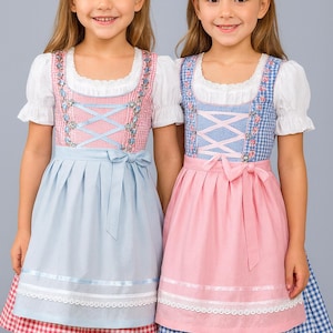May include: Two young girls wearing traditional German dirndl dresses. The dresses feature white blouses with ruffled sleeves, embroidered bodices, and full skirts. One dress is light blue with a pink bodice, and the other is pink with a blue bodice. Both have matching aprons.