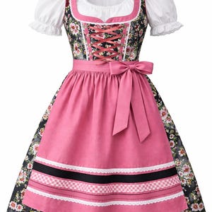 Op de afbeelding: Een traditionele dirndl-jurk met een witte blouse met pofmouwen, een bloemenlijfje en een roze rok met een bijpassende schort. De jurk heeft een roze lintcorset en een roze strik in de taille. De rok heeft witte kanten rand.