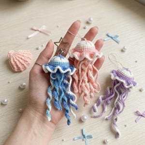 May include: Crocheted jellyfish keychains in blue, pink, and purple. Each jellyfish has a ruffled cap and long, curly tentacles. A crocheted seashell and pearl beads are also visible.