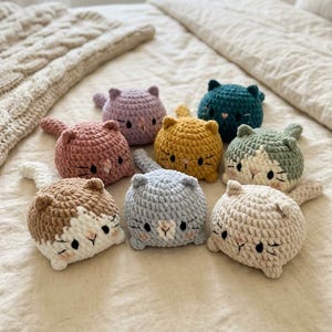 May include: A collection of eight small, round, crocheted cat plushies in various colours including lavender, teal, mustard yellow, dusty rose, light blue, cream, and brown-and-white patterns. Each cat features embroidered eyes, whiskers, and a tail.