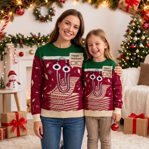 Puede incluir: Un suéter de punto rojo oscuro y verde con un gráfico de un personaje de clip con ojos grandes y un globo de texto que dice Happy Holidays! OK. El suéter tiene cuello redondo, puños y dobladillo acanalados en color crema claro.