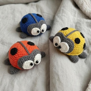 Puede incluir: Tres mariquitas hechas a mano con ganchillo en azul, naranja y amarillo. Cada bicho tiene manchas negras, detalles grises y ojos blancos grandes. Las mariquitas están sobre una tela gris claro.
