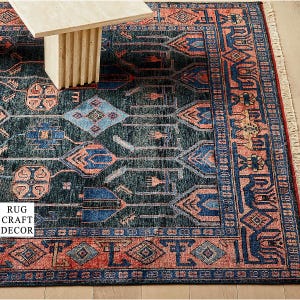 May include: A rectangular area rug featuring a dark blue background with intricate geometric patterns in shades of coral, light blue, and cream. The rug has a wide decorative border and a fringed edge. The text RUG CRAFT DECOR is visible.