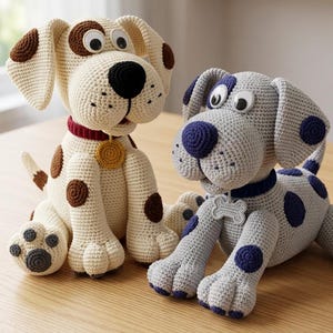 May include: Two handmade crochet dog stuffed animals. One is cream-colored with brown spots, a black nose, and a red collar. The other is gray with blue spots and a blue collar. Both have large, googly eyes and are sitting on a wooden table.
