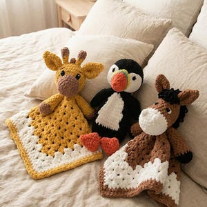 Puede incluir: Tres mantas de seguridad de animales de ganchillo: una jirafa, un pingüino y un caballo. La jirafa es amarilla con detalles marrones, el pingüino es blanco y negro con pico naranja, y el caballo es marrón y blanco.