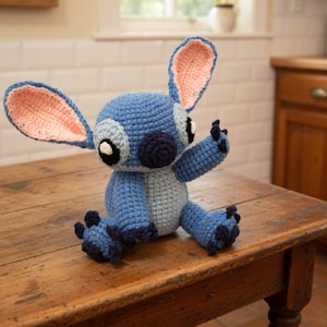 May include: A handmade blue crochet Stitch doll with large ears, black eyes, and a dark blue nose. The inside of the ears is a light pink. The doll is sitting on a wooden table.