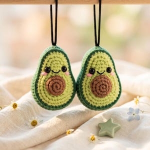 May include: Two crocheted avocado ornaments with smiling faces, black eyes, and pink cheeks hang from black cords. The avocados are green with brown centers. A green star and flowers are in the background.