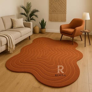 Op de afbeelding: Een roestkleurig, organisch gevormd vloerkleed met een golvend, concentrisch ontwerp. Het vloerkleed heeft de tekst "RUGSA" in de rechter benedenhoek. Het ligt op een houten vloer in een woonkamer.