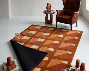 May include: A rectangular area rug featuring a geometric grid pattern in shades of orange, brown, and cream. The rug has a black backing visible where one corner is folded over. A logo with the letter R and the text RUGSA is printed on the rug.