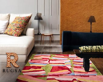 May include: A colorful area rug with a geometric design featuring red, pink, yellow, and blue stripes on a green background. The rug is in a living room setting with a white sofa, a dark blue sofa, and a wooden coffee table. The word "RUGSA" is in the image.