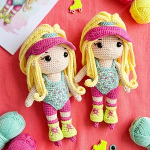 Può includere: Due bambole all'uncinetto fatte a mano con capelli biondi, visiere rosa e abiti a tema pattini a rotelle. Le bambole sono posizionate su una superficie rosa, circondate da gomitoli di lana di vari colori, uncinetti e materiali per l'artigianato.
