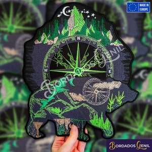 May include: Embroidered patch featuring a green and black bear with a compass and mountains in the background. The bear is standing on a green grassy area. The text "Bordados Genil" is visible in the lower right corner.