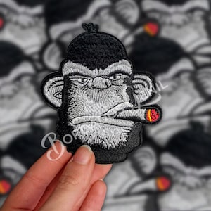 May include: Black and white embroidered patch of a monkey with a serious expression, smoking a red and orange cigar. The monkey has black fur and a white face.