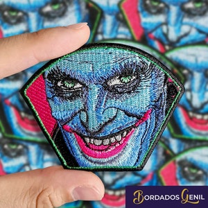 Può includere: Un toppa ricamata che rappresenta un mostro sorridente con labbra rosa e denti bianchi. La toppa è blu e verde, a forma di pentagono con un bordo nero.