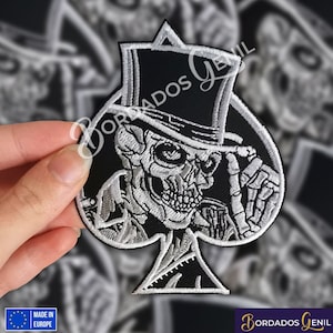 Może przedstawiać: Naszywkę haftowaną w kolorze czarno-białym przedstawiającą szkielet w cylindrze trzymającego kartę do gry. Karta to pik, a szkielet jest w pozie, jakby chciał zdjąć kapelusz. Naszywka jest otoczona innymi naszywkami.